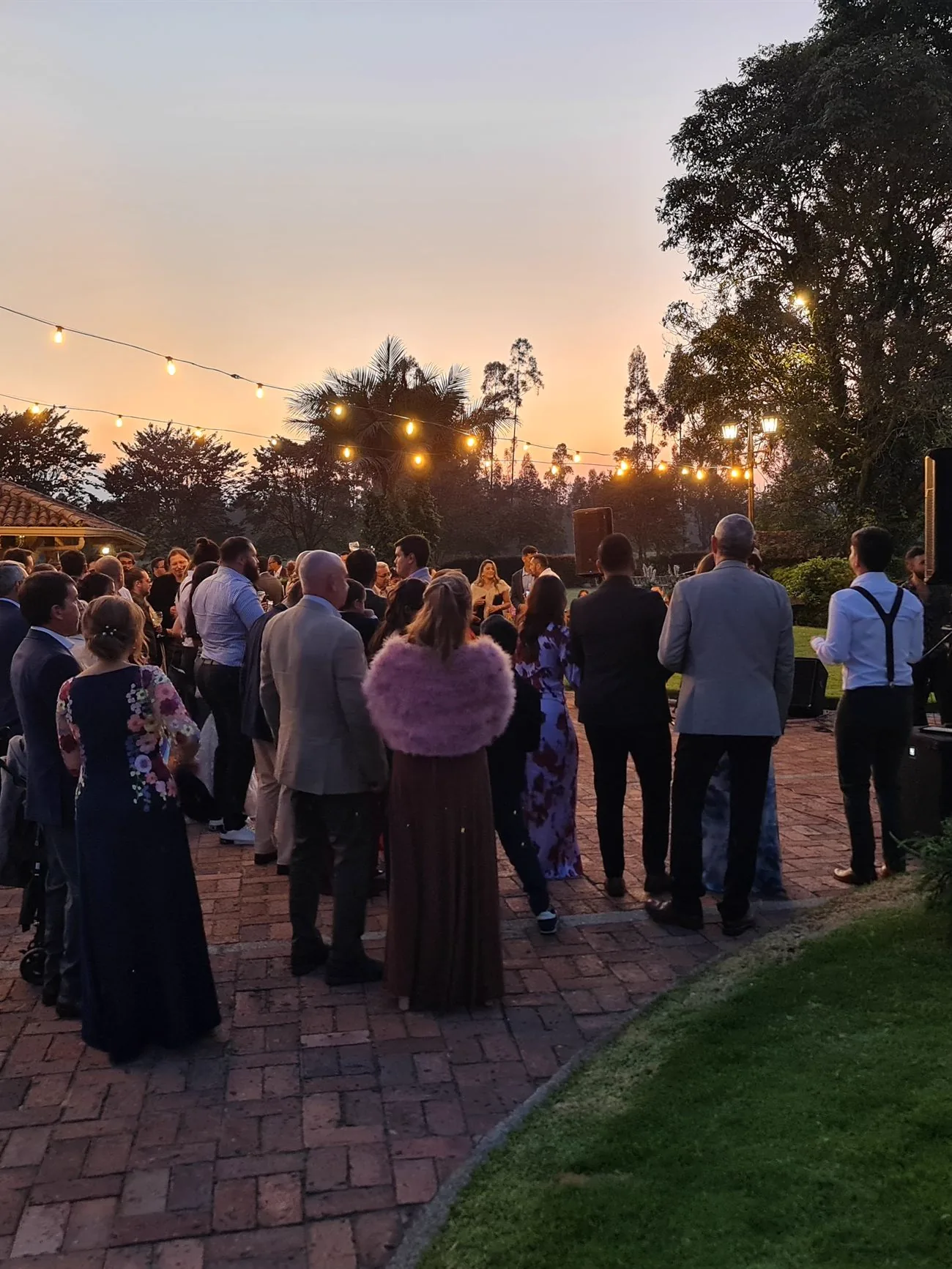 Cóctel de boda al atardecer: invitados de pie, banda en vivo y luces colgantes